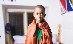 A kid standing on the bed with and adult shirt as a cape