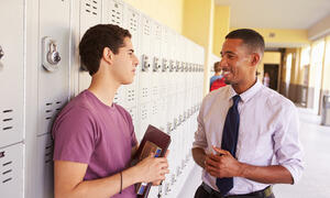 Student holding books speaking with adult in school hallway.