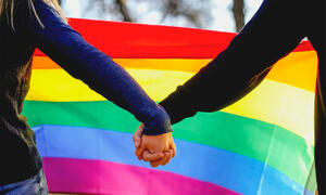 Two people holding hands in front of a rainbow flag.