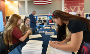 Two students, seated at a table covered in documents, facilitating a registration drive while another student fills out a sheet of paper across from them.