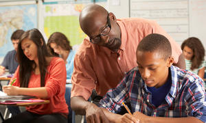 Teacher of color guiding student of color over their shoulder in a classroom setting.