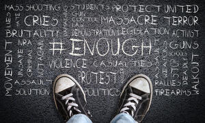 A child's feet with sneakers on them stand in front of words that include protect, students, mass shooting, cries, brutality, activism, walkout and more etched in chalk on the ground. 
