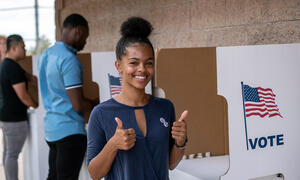 A young Black woman standing in front of a voting booth.