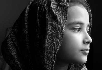 In a black and white photograph, a young girl wearing a head covering looks pensively off camera 