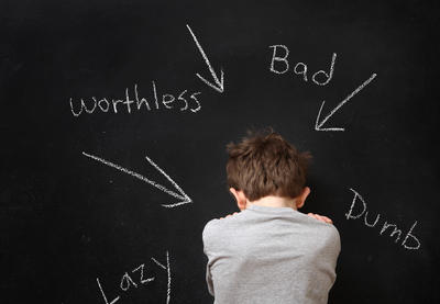 boy looking down shamefully from behind with words lazy worthless bad dumb written on chalkboard pointing to him