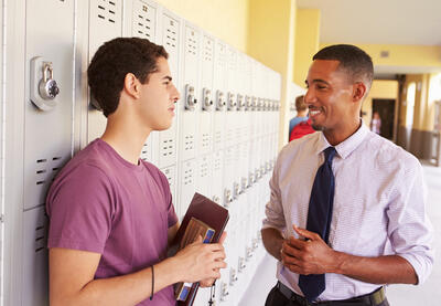 Student holding books speaking with adult in school hallway.