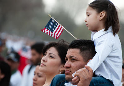 Young child holding American flag sitting on top of parent's shoulders.