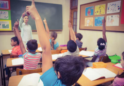 A classroom full of students raising their hands in front of their teacher.