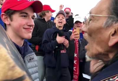 Young white student with "Make America Great Again" hat standing in front of elderly Native American man.