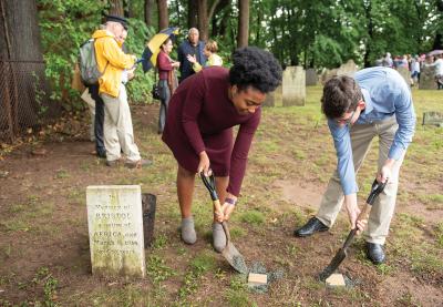 Two young people with spades digging near a burial marker.