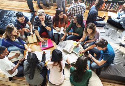Several older students in discussion with each other, gathered around two tables with open books on them.