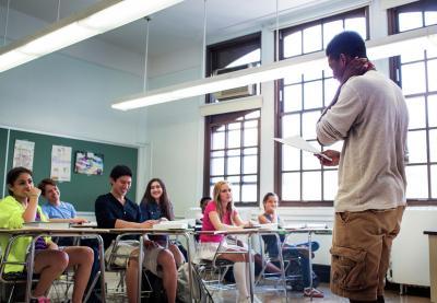 Student standing in front of classroom full of fellow students as they present something from a sheet of paper.