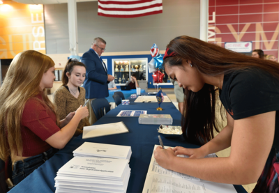 Two students, seated at a table covered in documents, facilitating a registration drive while another student fills out a sheet of paper across from them.