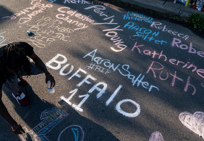 A teenager honors those who lost their lives in the Buffalo attack by writing their names in chalk on the street in front of a sidewalk memorial.