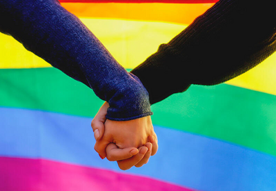 Two people hold hands with a rainbow flag in the background. 