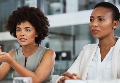 Two women sit at a table and one of the women is talking to someone across from her who is not in view. They are in a workshop setting.