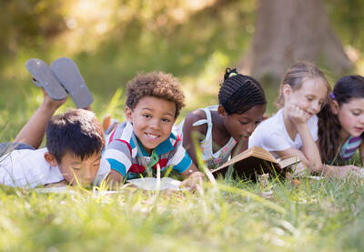 A group of children lie on their stomachs in the grass reading books in front of a tree on a sunny day. One child looks directly at the camera and smiles. 
