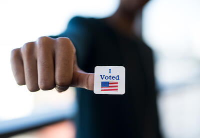 A young Black man's blurred image in the background with his hand and an "I voted" sticker and flag.