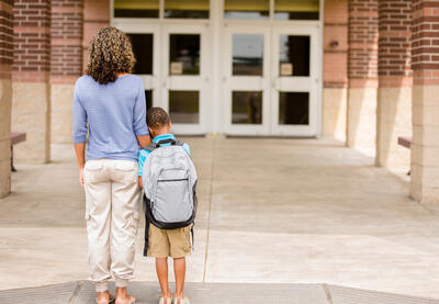 A mother supports her who leans against her as they stand in front of school doors.