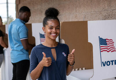 A young Black woman standing in front of a voting booth.