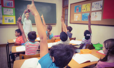 A classroom full of students raising their hands in front of their teacher.