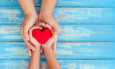 A pair of larger hands encircles a child's hands and in the child's hands is a red heart.