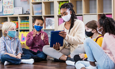 A teacher and a group of young students, all wearing masks, gather around in a half circle so they can look at images on an electronic tablet and listen during what appears to be story time. 