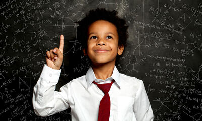 A smiling young black child stands in front of board filled with scientific formulas.