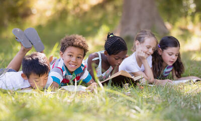 A group of children lie on their stomachs in the grass reading books in front of a tree on a sunny day. One child looks directly at the camera and smiles. 