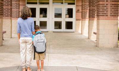 A mother supports her who leans against her as they stand in front of school doors.