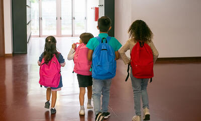 Four children with backpacks walking through a hallway towards a school's exit.