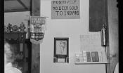 A black and white image of a wall behind a bar with signs that read "Positively No Beer Sold to Indians" and "God Bless America".