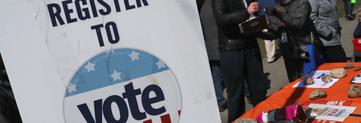 Piece of paper taped to a column that says "Register to Vote" with several people standing in the background interacting with each other.