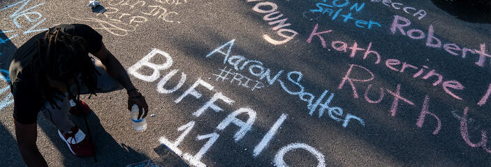 A teenager honors those who lost their lives in the Buffalo attack by writing their names in chalk on the street in front of a sidewalk memorial.