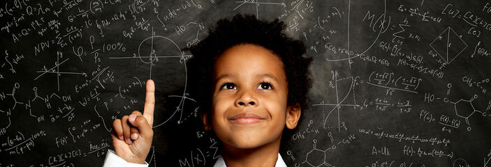 A smiling young black child stands in front of board filled with scientific formulas.