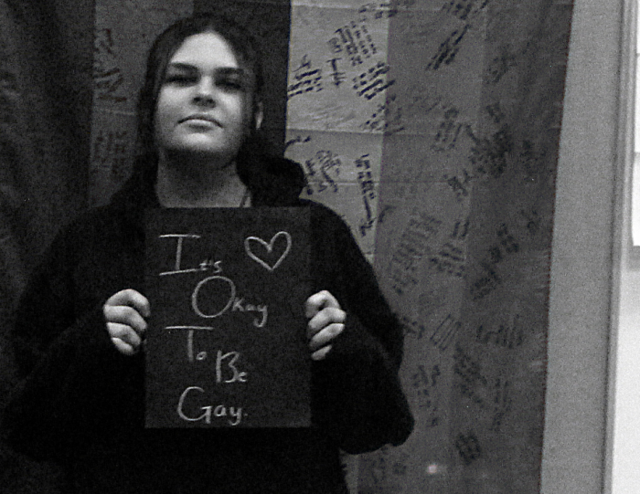 Young student holding sign that says "It's okay to be gay."