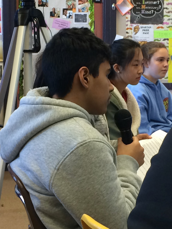 Young student in hoodie holding a microphone asking a question of the interviewee, who is offscreen.