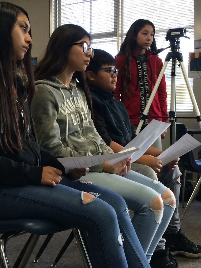 Several students holding sheets of paper with questions on them watching interviewee, who is offscreen and being filmed.