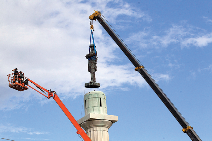 Photo of a crane removing a large statue of General Robert E. Lee from a tall pedestal, with workers supervising nearby.