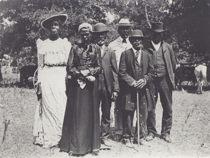 Six Black adults are stately dressed as they pose for this black and white photo taken against the backdrop of trees and a grassy areas where horse-drawn carriages await nearby.