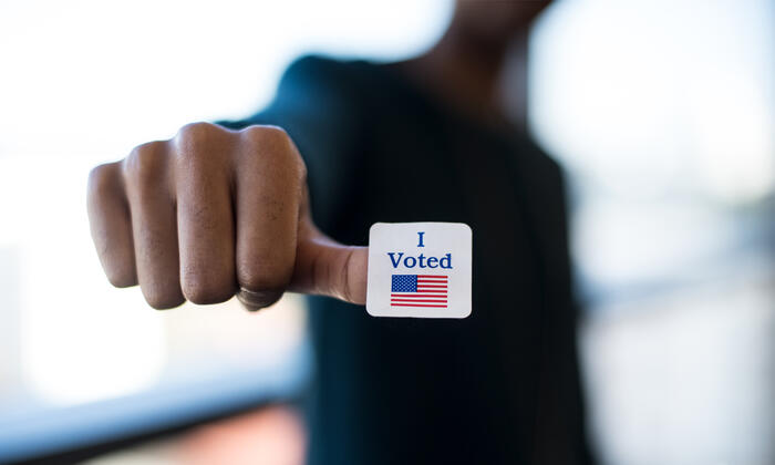 A young Black man's blurred image in the background with his hand and an "I voted" sticker and flag.