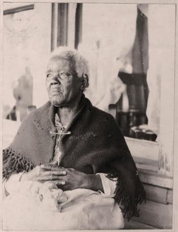 An elderly black woman sits outside a house